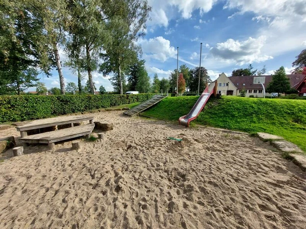 Der Spielplatz Alte Schule zeigt eine große Sandfläche mit einer Holz-Picknickbank. Eine Rutsche führt von einem grünen Hügel hinab. Im Hintergrund sind Bäume und ein Gebäude mit roten Dächern sichtbar. Der Himmel ist leicht bewölkt. Ideal für Kinder, um zu spielen und sich im Freien zu vergnügen.
