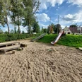 Spielplatz Alte Schule Der Spielplatz Alte Schule zeigt eine große Sandfläche mit einer Holz-Picknickbank. Eine Rutsche führt von einem grünen Hügel hinab. Im Hintergrund sind Bäume und ein Gebäude mit roten Dächern sichtbar. Der Himmel ist leicht bewölkt. Ideal für Kinder, um zu spielen und sich im Freien zu vergnügen.