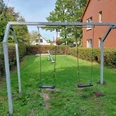 Spielplatz Torgelower Weg Zwei Schaukeln und eine Wippe stehen auf einer grünen Wiese, umrahmt von Bäumen und Hecken.