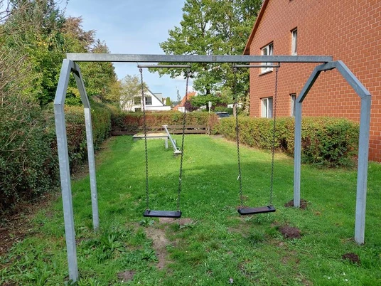 Spielplatz Torgelower Weg Zwei Schaukeln und eine Wippe stehen auf einer grünen Wiese, umrahmt von Bäumen und Hecken.