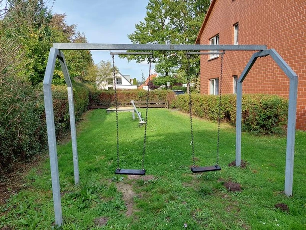 Spielplatz Torgelower Weg Zwei Schaukeln und eine Wippe stehen auf einer grünen Wiese, umrahmt von Bäumen und Hecken.