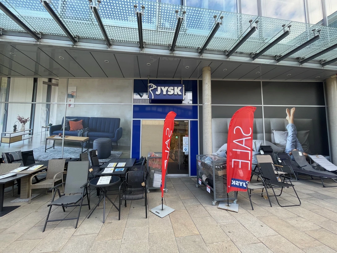 JYSK Stadtmitte, Wolfsburg.jpg Zu sehen ist die Eingangstür und die Schaufenster der Filiale JYSK. Die Schaufenster sind vollflächig mit Werbung beklebt. Vor dem Geschäft stehen verschiedene Gartenmöbel.