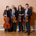 Vier Personen, zwei Frauen und zwei Männer, stehen nebeneinander in einem Raum mit einem Parkettboden und einer schlichten beigen Wand im Hintergrund. Jede Person hält ein Streichinstrument, darunter zwei Geigen, eine Bratsche und ein Cello. Alle tragen formelle, schwarze Kleidung.
