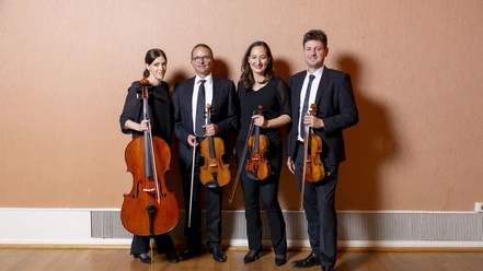 Vier Personen, zwei Frauen und zwei Männer, stehen nebeneinander in einem Raum mit einem Parkettboden und einer schlichten beigen Wand im Hintergrund. Jede Person hält ein Streichinstrument, darunter zwei Geigen, eine Bratsche und ein Cello. Alle tragen formelle, schwarze Kleidung.