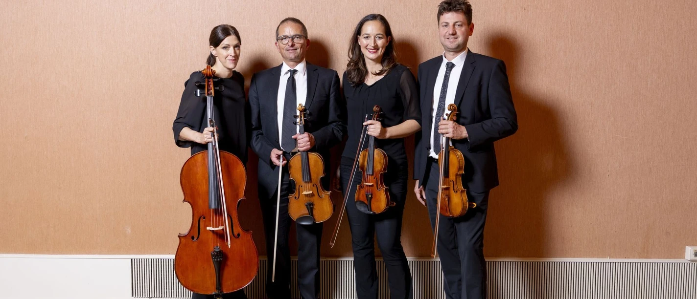 Vier Personen, zwei Frauen und zwei Männer, stehen nebeneinander in einem Raum mit einem Parkettboden und einer schlichten beigen Wand im Hintergrund. Jede Person hält ein Streichinstrument, darunter zwei Geigen, eine Bratsche und ein Cello. Alle tragen formelle, schwarze Kleidung.