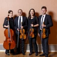 Vier im Couven_Chapelle Quartett (c) Chapelle Quartett.jpg Vier Personen, zwei Frauen und zwei Männer, stehen nebeneinander in einem Raum mit einem Parkettboden und einer schlichten beigen Wand im Hintergrund. Jede Person hält ein Streichinstrument, darunter zwei Geigen, eine Bratsche und ein Cello. Alle tragen formelle, schwarze Kleidung.