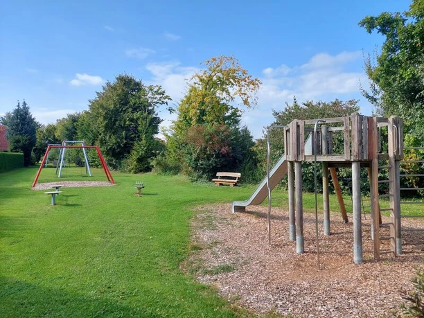 Ein Spielplatz mit Rutsche und Schaukeln auf grünem Rasen, umgeben von Bäumen unter blauem Himmel.