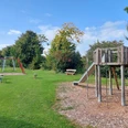 Spielplatz Im Ginsterfeld Ein Spielplatz mit Rutsche und Schaukeln auf grünem Rasen, umgeben von Bäumen unter blauem Himmel.