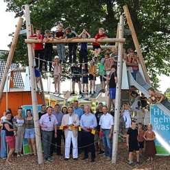 Spielplatz Alte Mühle Auf diesem Bild ist ein Spielplatz zu sehen. Kinder klettern fröhlich auf einem großen, hölzernen Klettergerüst, das mit Seilen und Rutschen ausgestattet ist. Erwachsene stehen unten und beobachten das Geschehen. Der Spielplatz ist unter freiem Himmel und von Bäumen umrahmt.