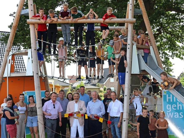 Spielplatz Alte Mühle Auf diesem Bild ist ein Spielplatz zu sehen. Kinder klettern fröhlich auf einem großen, hölzernen Klettergerüst, das mit Seilen und Rutschen ausgestattet ist. Erwachsene stehen unten und beobachten das Geschehen. Der Spielplatz ist unter freiem Himmel und von Bäumen umrahmt.