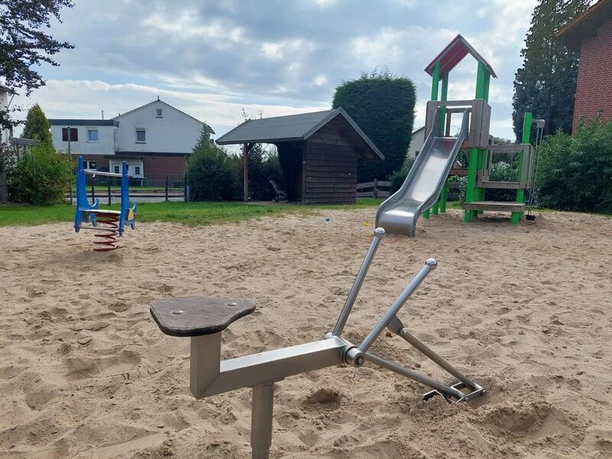 Das Bild zeigt einen Spielplatz mit einem hellen Sandboden. Im Vordergrund befindet sich ein Sandbagger aus Metall. Im Hintergrund steht ein Kletterturm mit einer Rutsche, farblich in Rot und Grün gehalten. Seitlich sind weitere Spielgeräte und ein kleines, hölzernes Gebäude zu erkennen, eingebettet in eine Wohnumgebung mit Häusern und Büschen im Hintergrund.