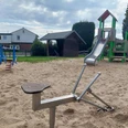 Spielplatz Hollenstraße Das Bild zeigt einen Spielplatz mit einem hellen Sandboden. Im Vordergrund befindet sich ein Sandbagger aus Metall. Im Hintergrund steht ein Kletterturm mit einer Rutsche, farblich in Rot und Grün gehalten. Seitlich sind weitere Spielgeräte und ein kleines, hölzernes Gebäude zu erkennen, eingebettet in eine Wohnumgebung mit Häusern und Büschen im Hintergrund.