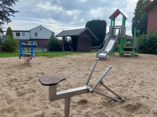 Spielplatz Hollenstraße Das Bild zeigt einen Spielplatz mit einem hellen Sandboden. Im Vordergrund befindet sich ein Sandbagger aus Metall. Im Hintergrund steht ein Kletterturm mit einer Rutsche, farblich in Rot und Grün gehalten. Seitlich sind weitere Spielgeräte und ein kleines, hölzernes Gebäude zu erkennen, eingebettet in eine Wohnumgebung mit Häusern und Büschen im Hintergrund.