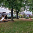 Spielplatz mit Holz-Klettergerüst, Rutsche und Schaukel, umgeben von Bäumen und Häusern im Hintergrund.