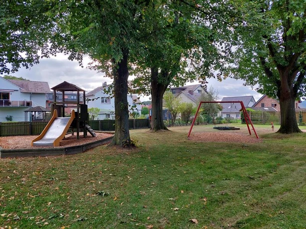 Spielplatz mit Holz-Klettergerüst, Rutsche und Schaukel, umgeben von Bäumen und Häusern im Hintergrund.