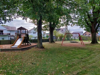 Spielplatz Neues Feld Spielplatz mit Holz-Klettergerüst, Rutsche und Schaukel, umgeben von Bäumen und Häusern im Hintergrund.