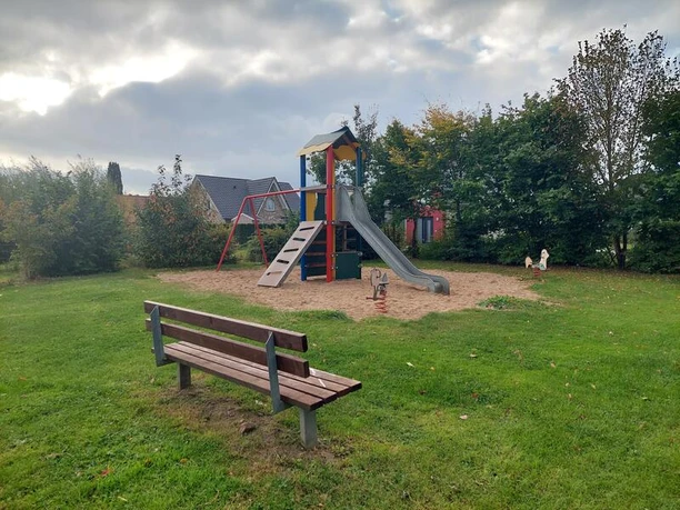 Spielplatz Röthenstraße Ein Spielplatz mit einer bunten Rutsche und Schaukel, umgeben von Bäumen unter einem bewölkten Himmel. Eine Holzbank steht auf einer Wiese.