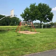 Spielplatz mit Basketballkorb, einer Schaukel, umgeben von grüner Landschaft und Bäumen.