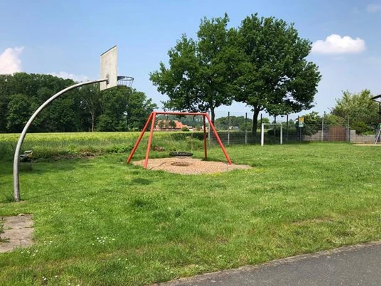 Spielplatz mit Basketballkorb, einer Schaukel, umgeben von grüner Landschaft und Bäumen.