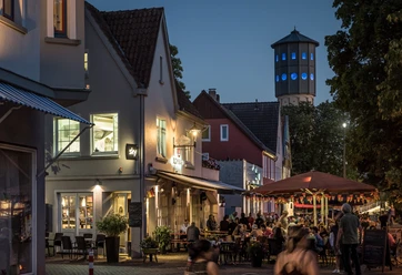 Kneipenmeile_Neu.jpg Abendliche Straßenszene mit beleuchtetem Wasserturm, Cafés, Restaurants und Flanieren in gemütlicher Atmosphäre.