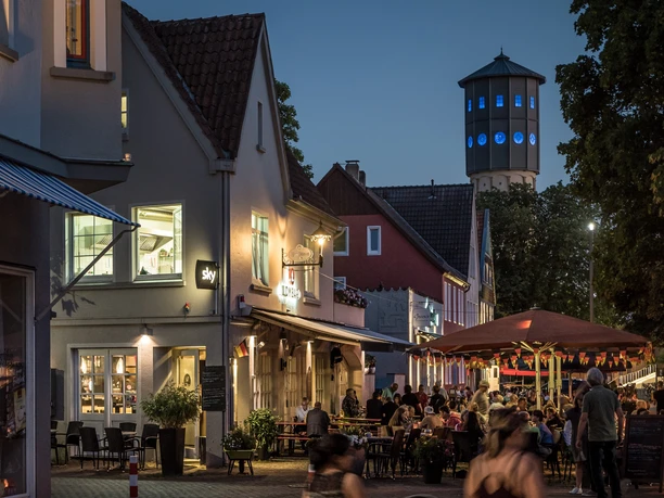 Kneipenmeile_Neu.jpg Abendliche Straßenszene mit beleuchtetem Wasserturm, Cafés, Restaurants und Flanieren in gemütlicher Atmosphäre.