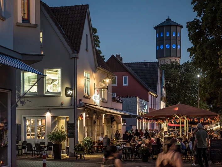 Kneipenmeile_Neu.jpg Abendliche Straßenszene mit beleuchtetem Wasserturm, Cafés, Restaurants und Flanieren in gemütlicher Atmosphäre.
