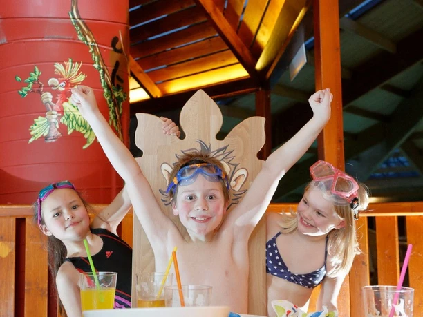 Drei fröhliche Kinder mit Schwimmbrillen heben die Arme, umgeben von bunten Farben und Getränken.