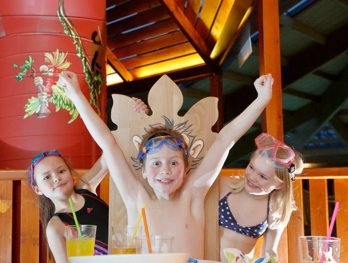 Drei fröhliche Kinder mit Schwimmbrillen heben die Arme, umgeben von bunten Farben und Getränken.