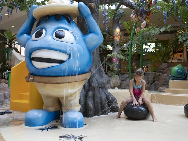 Blaues Wassermaskottchen neben spielendem Kind in einem Erlebnisbad mit Wasserfontänen und Pflanzen.