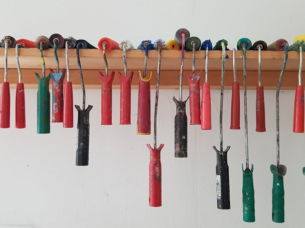 20220204_121436.jpg Verschiedenfarbige Malwerkzeuge hängen geordnet an einer Holzleiste an einer weißen Wand.