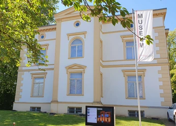 Museum Sommer I.jpg Ein cremefarbiges, zweigeschossiges Gebäude mit symmetrischer Fassade und Museumsbanner davor.