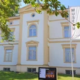 Museum Sommer I.jpg Ein cremefarbiges, zweigeschossiges Gebäude mit symmetrischer Fassade und Museumsbanner davor.