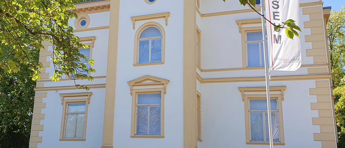 Museum Sommer I.jpg Ein cremefarbiges, zweigeschossiges Gebäude mit symmetrischer Fassade und Museumsbanner davor.