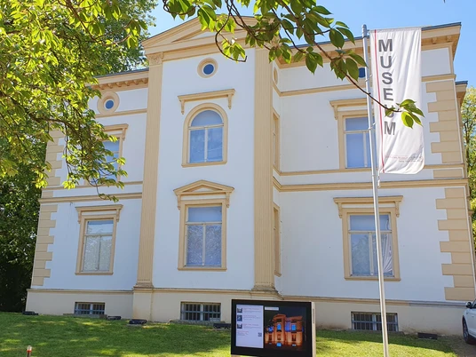 Museum Sommer I.jpg Ein cremefarbiges, zweigeschossiges Gebäude mit symmetrischer Fassade und Museumsbanner davor.