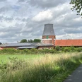 Wesermühle GbR Backsteinmühle mit zylinderförmigem Turm und roten Dächern vor bewölktem Himmel, umgeben von Wiesen.