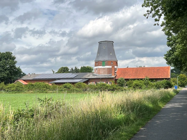 Wesermühle GbR Backsteinmühle mit zylinderförmigem Turm und roten Dächern vor bewölktem Himmel, umgeben von Wiesen.