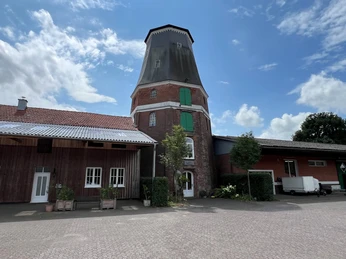 Wesermühle GbR Historische Windmühle in Backsteinoptik mit grünen Türen, umgeben von rustikalen Gebäuden.