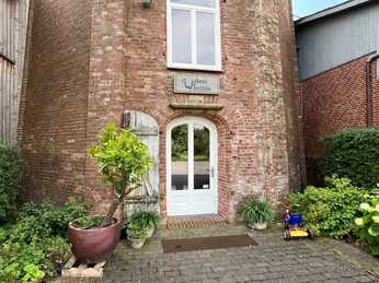 Wesermühle GbR Eingang einer alten Backsteinmühle mit großen Holztüren, umgeben von Pflanzen und einem Kinderspielzeug.