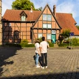 herford-altstadt-teutoburger-wald-tourismus-h-tornow-015-jpg.jpg Zwei Personen stehen vor einem historischen Fachwerkhaus mit rotem Ziegeldach in der Altstadt Herfords.
