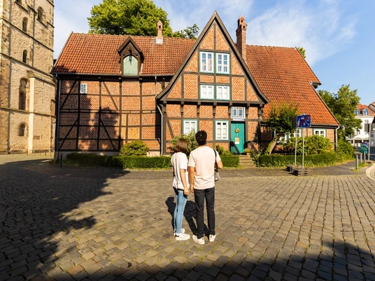 herford-altstadt-teutoburger-wald-tourismus-h-tornow-015-jpg.jpg Zwei Personen stehen vor einem historischen Fachwerkhaus mit rotem Ziegeldach in der Altstadt Herfords.