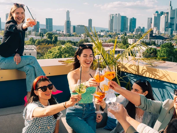 The Blasky Rooftop Freunde stoßen mit Getränken auf Dachterrasse mit Skyline an.