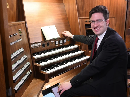 7_14August_cWolfgangSchmidt.JPG Ein Mann in Anzug und Krawatte sitzt lächelnd an einer großen Holzorgel mit mehreren Tastaturen und Knöpfen, ein aufgeschlagenes Buch liegt darauf.A man in a suit and tie sits smiling at a large wooden organ with several keyboards and buttons, an open book lying on top.Muž v obleku a kravatě sedí s úsměvem u velkých dřevěných varhan s několika klávesami a tlačítky, na kterých leží otevřená kniha.Mężczyzna w garniturze i krawacie siedzi uśmiechnięty przy dużych drewnianych organach z kilkoma klawiaturami i przyciskami, a na górze leży otwarta książka.Een man in pak met stropdas zit glimlachend aan een groot houten orgel met verschillende toetsenborden en knoppen, een open boek ligt bovenop.Un uomo in giacca e cravatta siede sorridente a un grande organo di legno con diverse tastiere e pulsanti, con un libro aperto sopra.