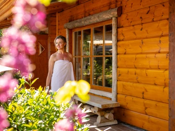 Herford-2025-117-©Teutoburger Wald - Pro Herford GmbH - D. Ketz.jpg Eine Frau steht in ein Handtuch gewickelt vor einem hölzernen Blockhaus, umgeben von blühenden Pflanzen.