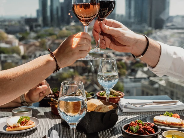 OBEN im Melia-Frankfurt-City Zwei Personen stoßen mit Wein an, Tapas auf gedecktem Tisch am Fenster.