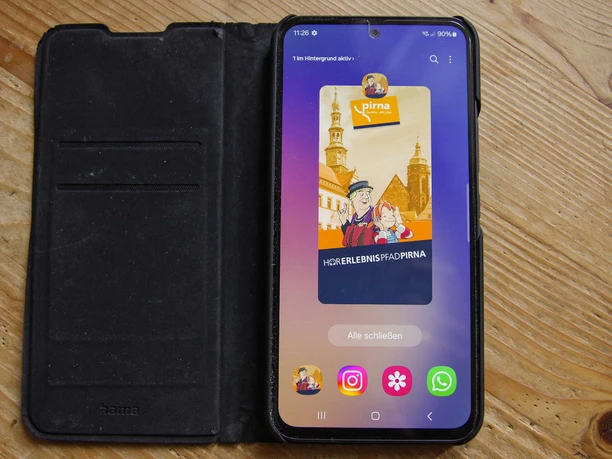 Smartphone in een zwart hoesje op een houten tafel toont een app met de titel "Hörerlebnispfad Pirna" en een illustratie van twee mensen voor gebouwen.