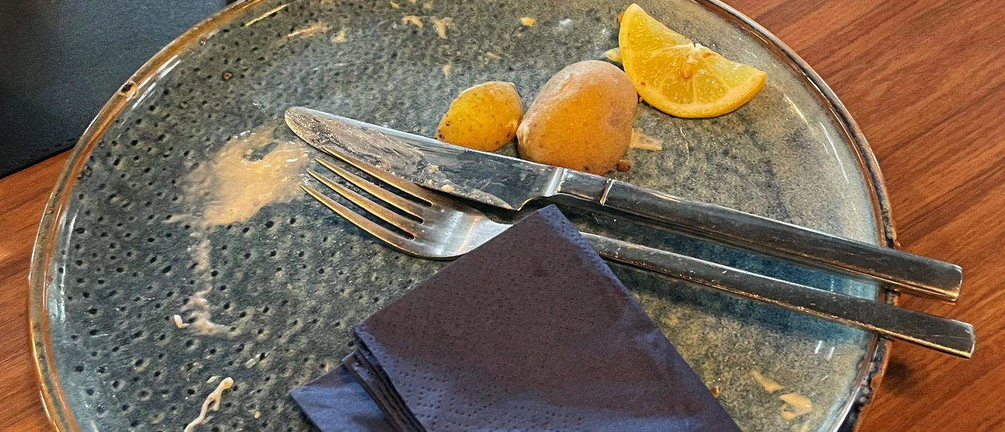 Aufgegessen, war lecker!-Platzhalterbild.jpg Leerer Teller mit Messer, Gabel, zwei Kartoffeln und Zitronenscheibe, daneben ein Wasserglas.