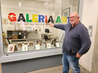 Ein Mann zeigt lächelnd auf bunte "GALERIA ART" Buchstaben in einem Schaufenster voller Kunst.