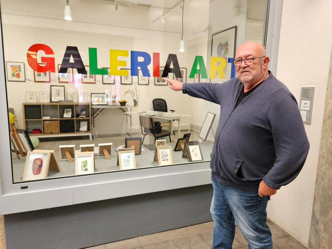 Ein Mann zeigt lächelnd auf bunte "GALERIA ART" Buchstaben in einem Schaufenster voller Kunst.