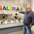 Ein Mann zeigt lächelnd auf bunte "GALERIA ART" Buchstaben in einem Schaufenster voller Kunst.
