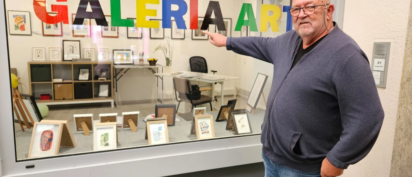 Galeria Arte_Gerhard Pollheide_1000011663.jpg Ein Mann zeigt lächelnd auf bunte "GALERIA ART" Buchstaben in einem Schaufenster voller Kunst.